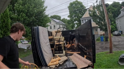 Crew loading a mattress, chairs, and rolled rug into a dump trailer on a residential street